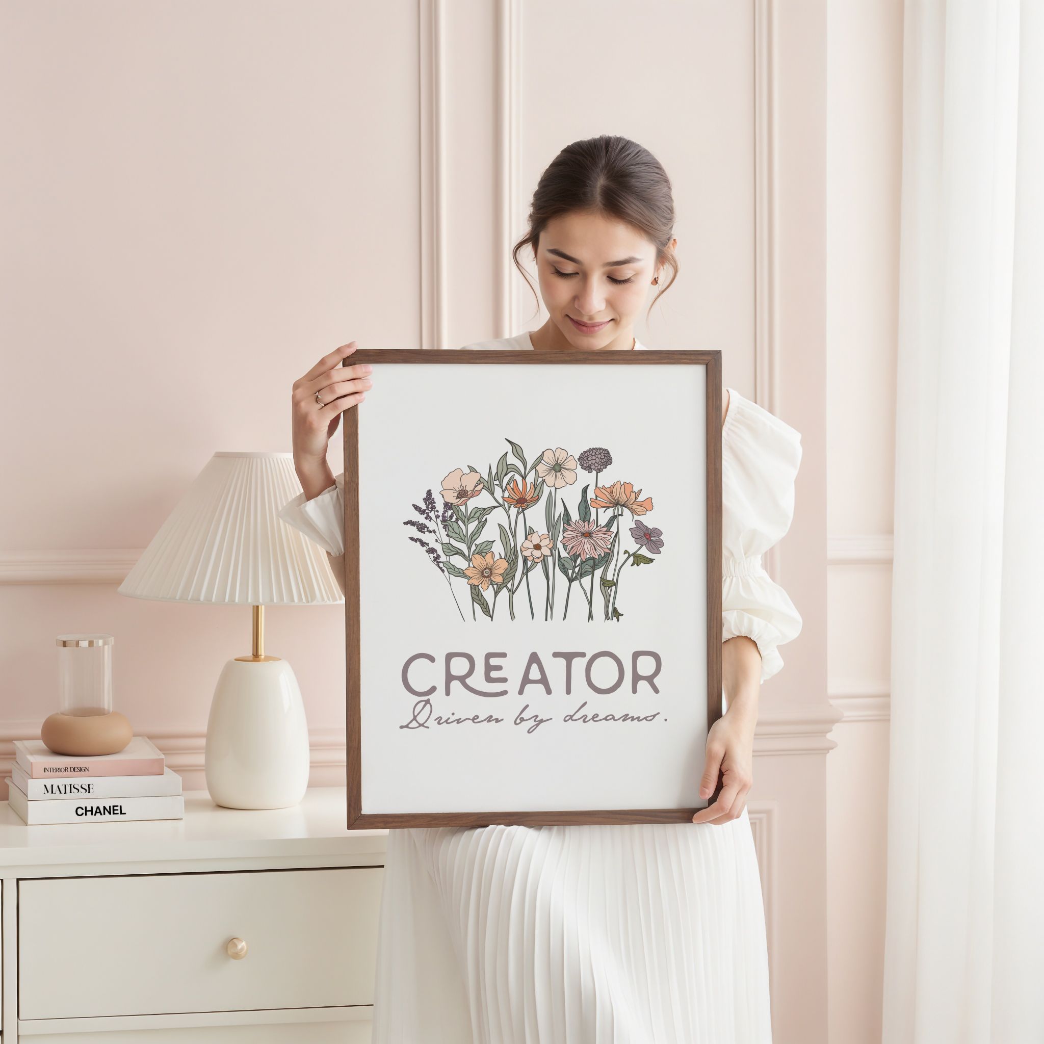 Woman holding framed artwork with floral design and text 'Creator' in a soft pink bedroom with a lamp and books.