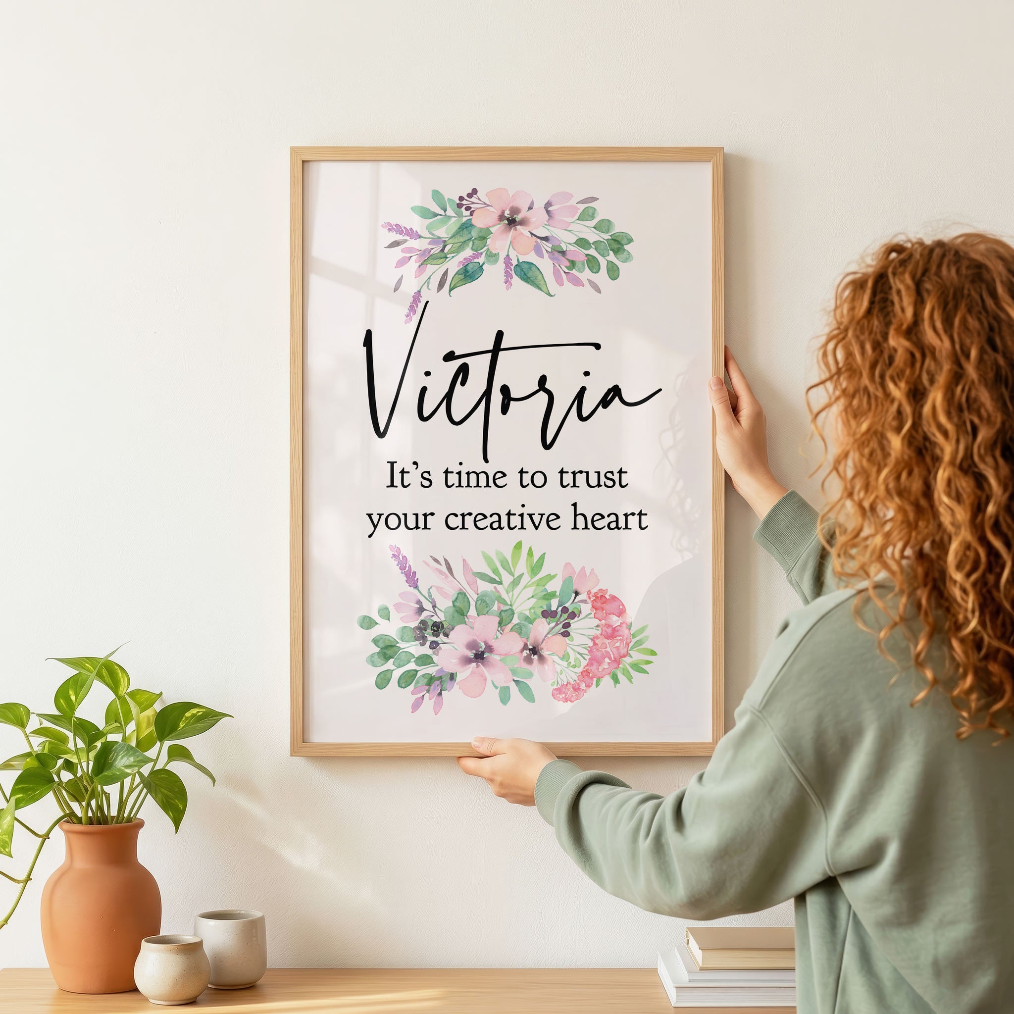 Red-haired woman hanging a framed print with floral design and text on a wall.