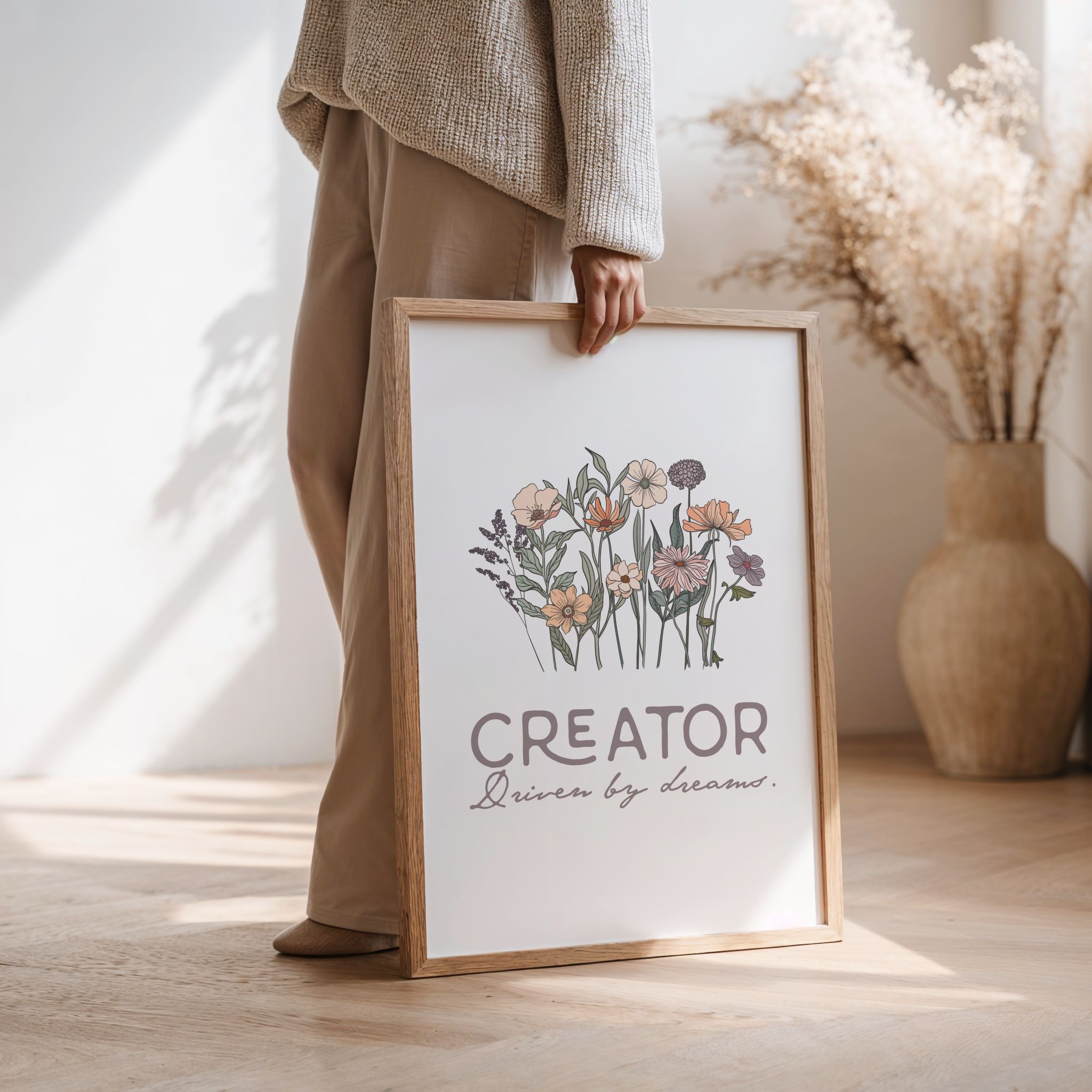 Woman holding a framed print with floral design and text 'Creator Driven by dreams' in a home setting