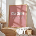 Framed artwork with 'Cha-Ching is my love language' text on a pink background, placed on a wooden chest.