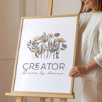 Smiling woman holding framed artwork with floral design and text 'Creator' on an easel.