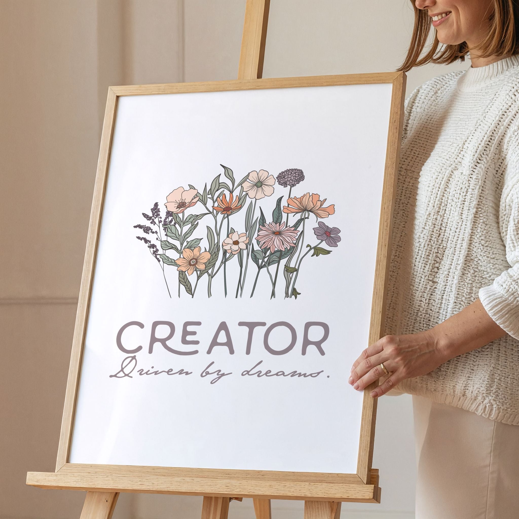 Smiling woman holding framed artwork with floral design and text 'Creator' on an easel.