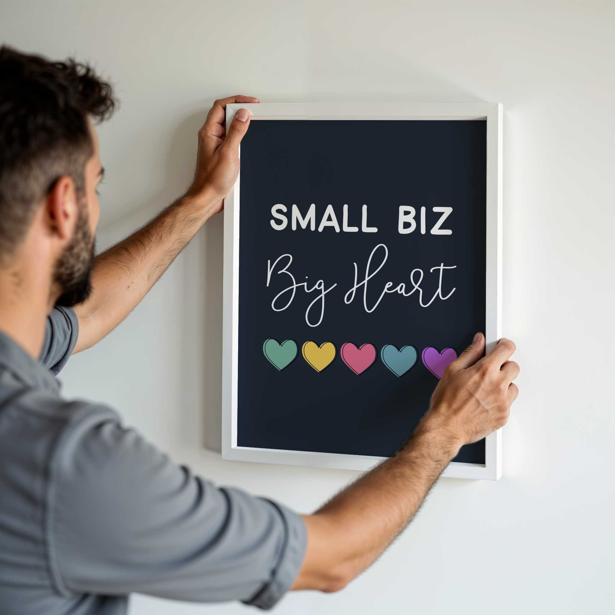 Man hanging a framed sign with 'Small Biz Big Heart' text and colorful hearts on a white wall.