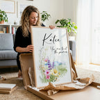Woman holding a framed print with floral design and text in a home setting